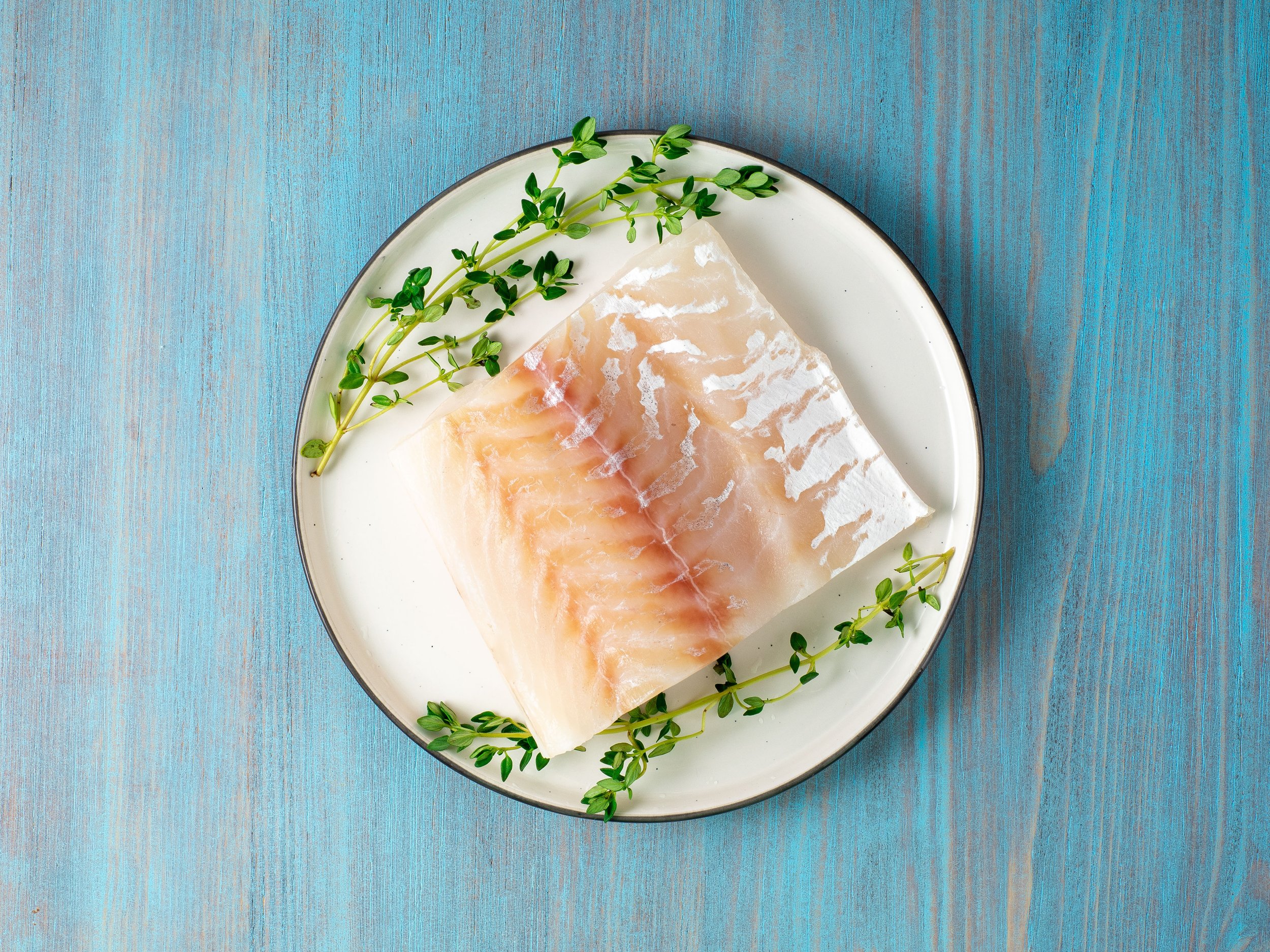 piece of raw cod fish fillet on plate on blue wooden table, top view Fresh fish fillet garnished with thyme on a white plate, set against a blue wooden background, showcasing a healthy and appetizing seafood dish.